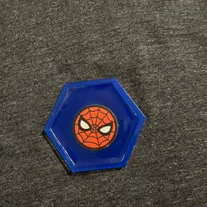 Puede incluir: Un posavasos hexagonal azul con el logotipo de Spider-Man. El logotipo es un dise&ntilde;o de telara&ntilde;a roja con ojos blancos sobre un fondo rojo. El posavasos est&aacute; hecho de un material brillante.