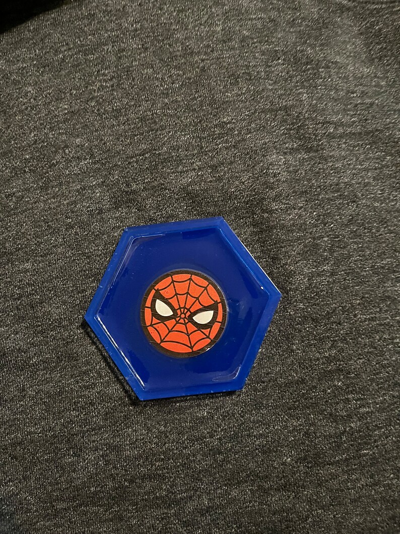Puede incluir: Un posavasos hexagonal azul con el logotipo de Spider-Man. El logotipo es un dise&ntilde;o de telara&ntilde;a roja con ojos blancos sobre un fondo rojo. El posavasos est&aacute; hecho de un material brillante.