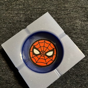 Puede incluir: Un cenicero cuadrado con interior azul y exterior azul claro. El centro presenta un gr&aacute;fico de Spider-Man con un dise&ntilde;o de telara&ntilde;a roja, ojos blancos y un contorno negro.