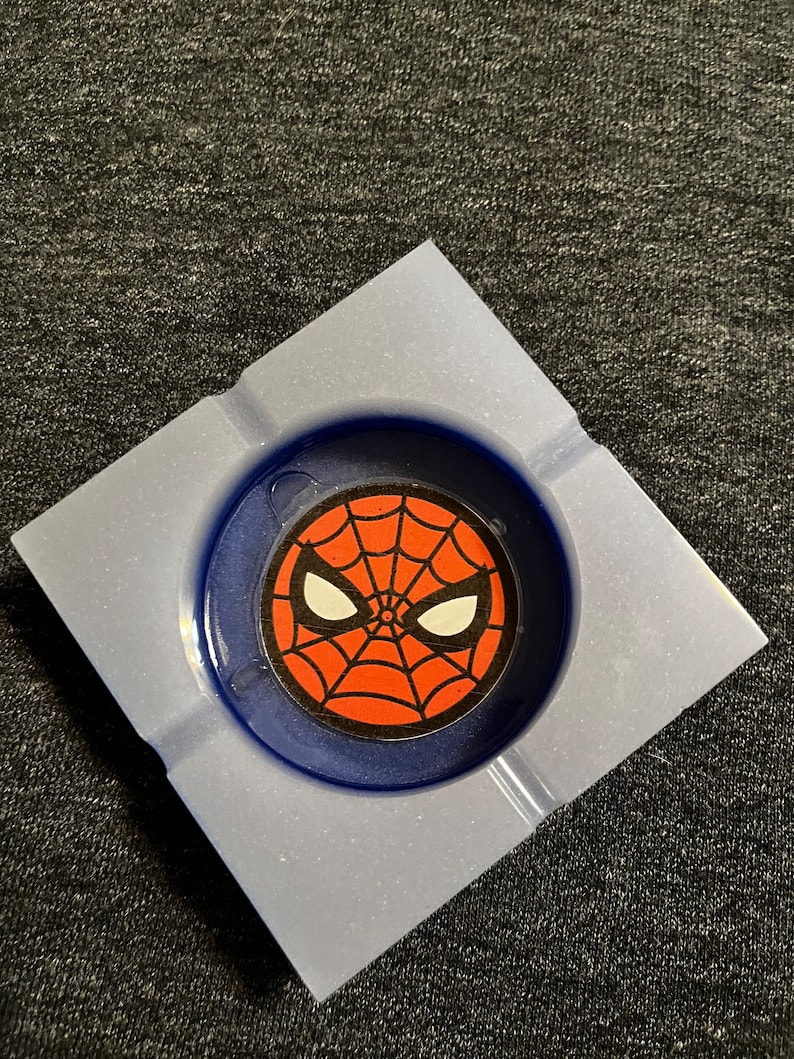 Puede incluir: Un cenicero cuadrado con interior azul y exterior azul claro. El centro presenta un gr&aacute;fico de Spider-Man con un dise&ntilde;o de telara&ntilde;a roja, ojos blancos y un contorno negro.