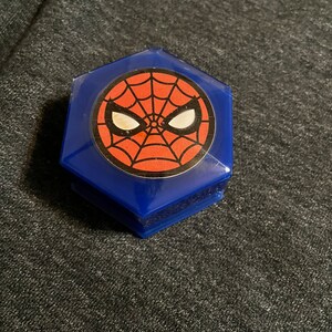 Puede incluir: Una caja hexagonal azul con un dise&ntilde;o de Spider-Man. El dise&ntilde;o incluye un fondo de telara&ntilde;a rojo con ojos blancos y un contorno negro. La caja est&aacute; hecha de pl&aacute;stico brillante y transparente.