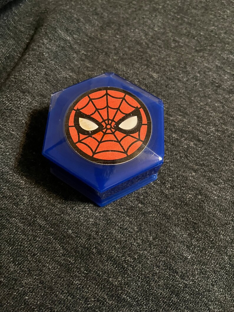 Puede incluir: Una caja hexagonal azul con un dise&ntilde;o de Spider-Man. El dise&ntilde;o incluye un fondo de telara&ntilde;a rojo con ojos blancos y un contorno negro. La caja est&aacute; hecha de pl&aacute;stico brillante y transparente.
