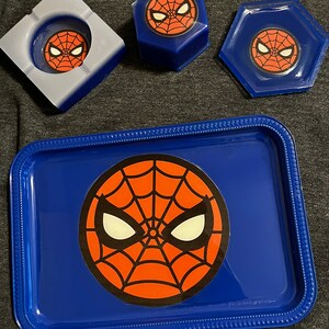 Puede incluir: Una bandeja azul, un cenicero y dos recipientes con el dise&ntilde;o de la m&aacute;scara de Spider-Man. La bandeja tiene un borde elevado y un gran gr&aacute;fico de la m&aacute;scara. El cenicero es cuadrado y los recipientes son hexagonales.