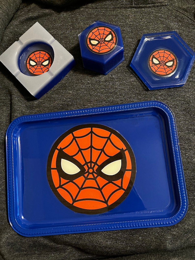 Puede incluir: Una bandeja azul, un cenicero y dos recipientes con el dise&ntilde;o de la m&aacute;scara de Spider-Man. La bandeja tiene un borde elevado y un gran gr&aacute;fico de la m&aacute;scara. El cenicero es cuadrado y los recipientes son hexagonales.