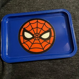 Puede incluir: Una bandeja met&aacute;lica azul con un dise&ntilde;o de Spider-Man. La bandeja tiene forma rectangular redondeada con un borde elevado. El gr&aacute;fico de Spider-Man est&aacute; en el centro, con un dise&ntilde;o de telara&ntilde;a roja y ojos blancos.