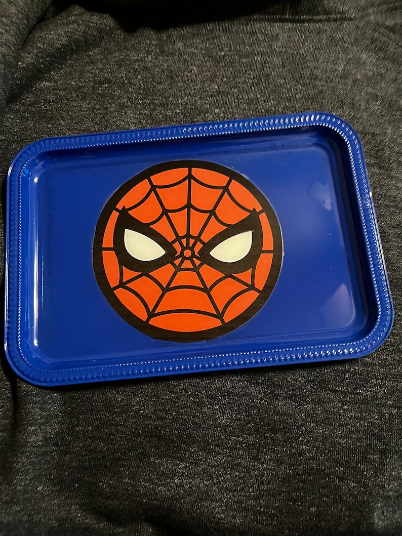 Puede incluir: Una bandeja met&aacute;lica azul con un dise&ntilde;o de Spider-Man. La bandeja tiene forma rectangular redondeada con un borde elevado. El gr&aacute;fico de Spider-Man est&aacute; en el centro, con un dise&ntilde;o de telara&ntilde;a roja y ojos blancos.