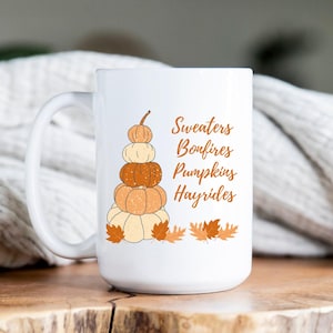 Könnte beinhalten: Weiße Keramik-Kaffeetasse mit einer Grafik eines Stapels Kürbisse und dem Text "Sweaters Bonfires Pumpkins Hayrides".