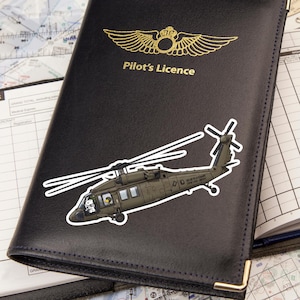 May include: A black leather Pilot's Licence cover featuring a gold embossed winged emblem and a sticker of a military helicopter with a cartoon character in the cockpit. The cover has gold-toned metal corner protectors and a smooth texture.