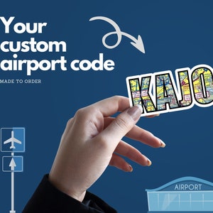 May include: A sticker with the airport code "KAJO" in a map-like design. The sticker is being held by a hand in front of a blue background with a small airport illustration.
