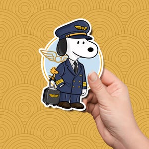 May include: A die-cut sticker featuring a white dog dressed in a navy blue pilot uniform with a matching cap, gold accents, and a black tie. A small yellow bird stands on a black rolling suitcase next to the dog, set against a light blue circle.
