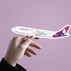 May include: A white and purple Hawaiian Airlines airplane sticker. The sticker features the airline's logo and a design on the tail. The airplane is held up by a hand against a light purple background.