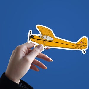 May include: A yellow airplane sticker with a red and white tail. The sticker is being held by a hand against a blue background.