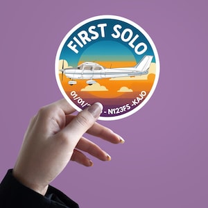 May include: A round sticker with a white border and a graphic of a small airplane. The sticker reads "FIRST SOLO" in blue text, with the plane set against a sunset background. The text "01/01/" and "-N123FS-KAJO" are also visible.
