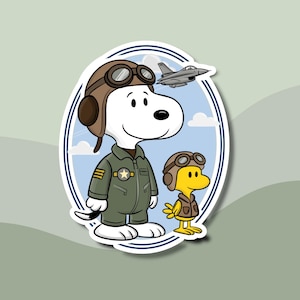 May include: A sticker featuring Snoopy and Woodstock in aviator gear. Snoopy is wearing a green flight suit and a brown leather helmet with goggles. Woodstock is in a similar outfit. A gray jet flies above them against a blue sky.