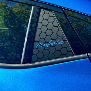 May include: Close-up of a blue car window with a black honeycomb pattern and the word "Rafale" in blue script. The car's blue paint has a metallic sheen, reflecting the sunlight.