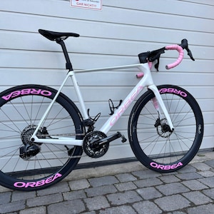 Può includere: Una bici da strada bianca con ruote nere e dettagli rosa. La bici presenta il marchio "ORBEA" in rosa sulle ruote e sul telaio. Il manubrio è avvolto con nastro rosa. La bici è parcheggiata davanti a una porta del garage.