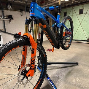 以下が含まれることがあります： オレンジのアクセントが付いた青と黒の自転車が、黒い自転車スタンドの上に置かれています。自転車はフォックスサスペンションフォークとフレームに「CUBE」のロゴが付いています。自転車はガレージにあります。