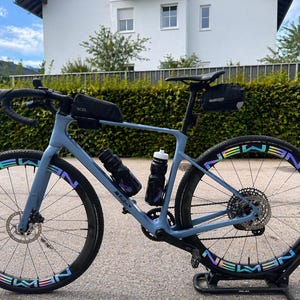 Könnte beinhalten: Ein hellblaues Rennrad mit schwarzen Reifen und schillernden Buchstaben auf den Felgen. Das Fahrrad hat einen schwarzen Sattel, Lenker und Flaschenhalter. Es steht auf einem schwarzen Ständer, mit einer grünen Hecke und einem weißen Haus im Hintergrund.