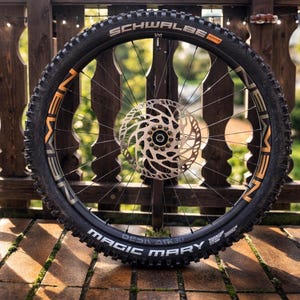May include: A black mountain bike wheel with a silver disc brake rotor. The tire has the text "SCHWALBE" and "MAGIC MARY" in white. The wheel is set against a wooden railing.
