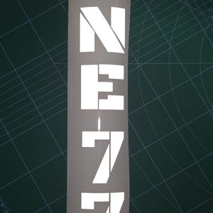 Könnte beinhalten: Ein vertikaler weißer Vinyl-Aufkleber mit dem Text ONE77 in einer fetten, blockartigen Schablonenschrift. Die Buchstaben und Zahlen sind in einer einzigen Spalte auf einer weißen Trägerfolie angeordnet und weisen scharfe, kantige Formen auf.