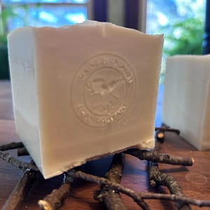 Op de afbeelding: Een vierkant blok gebroken witte zeep met een cirkelvormige reliëfstempel met de tekst Maine Highland Soap Company en een afbeelding van een eland in het midden. De zeep rust op een klein arrangement van donkerbruine takjes.