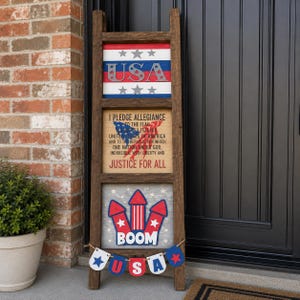 May include: A tall, dark brown wooden ladder display featuring three square signs. The top sign shows red, white, and blue stripes with the text USA. The middle sign displays the Pledge of Allegiance. The bottom sign features red rockets and the word BOOM, with a small bunting banner hanging below.