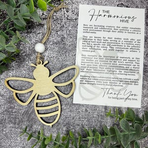 May include: A wooden bee ornament with a white bead hanging from a string. The ornament is in front of a card with the text "The Harmonious Hive" and a description of the importance of teamwork and collaboration. The card also says "Thank You for being part of my hive."