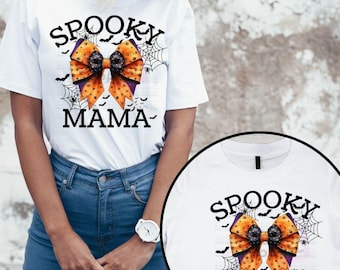 Può includere: Una t-shirt bianca a maniche corte con la grafica di un fiocco a pois arancione e nero con ragnatele e pipistrelli. Il testo recita SPOOKY MAMA. Un inserto circolare mostra un design abbinato su una t-shirt con il testo SPOOKY MINI.