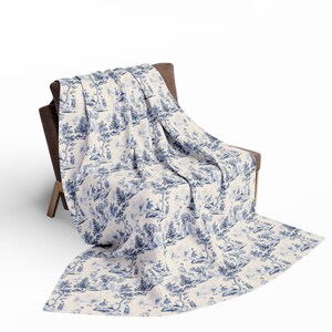 May include: A rectangular throw blanket featuring a blue and white toile pattern with detailed illustrations of trees, foliage, and small figures in outdoor settings. The fabric is draped over a brown chair, displaying a repeating pastoral scene.