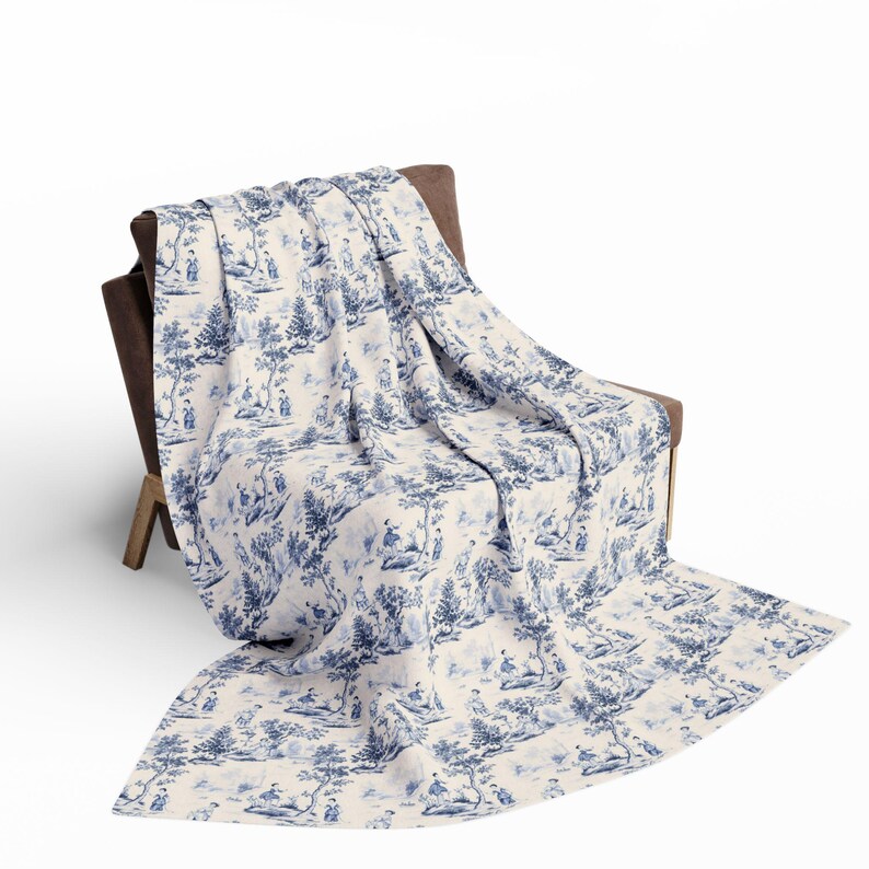 May include: A rectangular throw blanket featuring a blue and white toile pattern with detailed illustrations of trees, foliage, and small figures in outdoor settings. The fabric is draped over a brown chair, displaying a repeating pastoral scene.