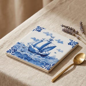 May include: Square ceramic coaster with a blue and white nautical design featuring a sailboat on the ocean. The coaster has floral border details and is accompanied by a gold spoon and lavender sprigs.