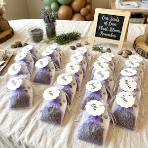 May include: A collection of small, clear bags filled with dried lavender, each tied with a purple ribbon and a white tag with a name. A small chalkboard sign reads "Our Seeds of Love: Plant, Bloom, Remember."