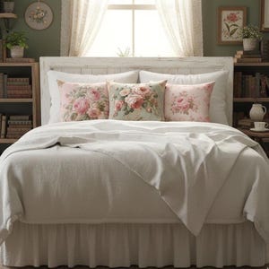 May include: A bed with a white headboard and white bedding. Three decorative throw cushions with floral patterns in pink and green are arranged on the bed. The room has a vintage aesthetic, with framed artwork and lace curtains.