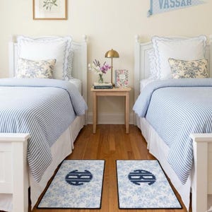 May include: Two twin beds with white headboards and blue and white striped bedding. Floral patterned throw pillows and matching monogrammed rugs in front of each bed. A small table with a lamp and flowers sits between the beds.