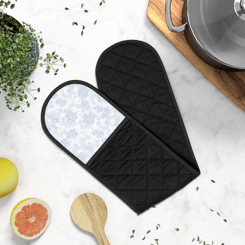 Puede incluir: Una manopla doble para horno con tela acolchada negra en el exterior y un estampado floral azul claro en el interior. La manopla tiene un dise&ntilde;o largo y conectado con dos bolsillos para las manos, acabado con ribetes negros en los bordes.
