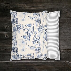 May include: A rectangular throw pillow featuring a cream-colored fabric with a blue toile de Jouy pattern depicting figures in a landscape on one side, and a grey and white horizontal striped pattern on the other side of the pillow cover.