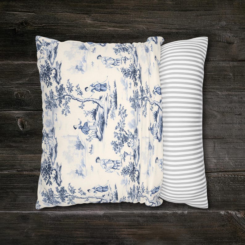 May include: A rectangular throw pillow featuring a cream-colored fabric with a blue toile de Jouy pattern depicting figures in a landscape on one side, and a grey and white horizontal striped pattern on the other side of the pillow cover.