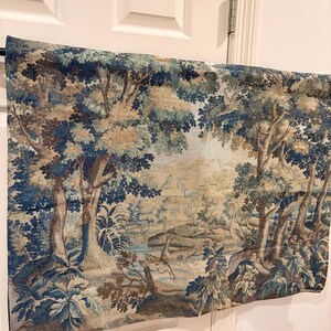 May include: A tapestry depicting a serene forest scene with trees, a river, and wildlife. The artwork features a color palette of blues, greens, and browns, creating a vintage aesthetic. The tapestry is hanging on a white door.