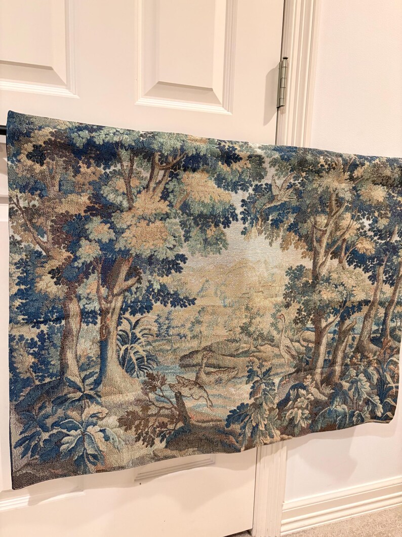 May include: A tapestry depicting a serene forest scene with trees, a river, and wildlife. The artwork features a color palette of blues, greens, and browns, creating a vintage aesthetic. The tapestry is hanging on a white door.