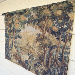 May include: A tapestry depicting a forest scene with trees, a river, and birds. The tapestry features shades of blue, green, and brown. It is hanging on a black rod against a white wall.