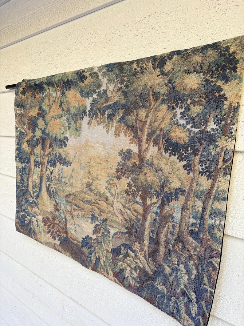 May include: A tapestry depicting a forest scene with trees, a river, and birds. The tapestry features shades of blue, green, and brown. It is hanging on a black rod against a white wall.