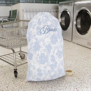 May include: A white laundry bag featuring a light blue floral pattern and the name Brooke written in a cursive script. The bag has a drawstring closure and is made of fabric, designed for holding clothing items in a laundry room setting.