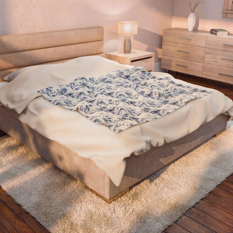 May include: A rectangular throw blanket featuring a blue and white toile pattern with intricate pastoral scenes, draped over a beige upholstered bed frame. The blanket displays detailed illustrations of landscapes and figures in a classic monochromatic style.