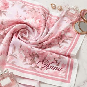 May include: A pink floral scarf with the name "Anna" in cursive script. The scarf features a repeating pattern of pink flowers and a white border with pink stripes. Accessories like earrings, perfume, and a compact mirror are also visible.