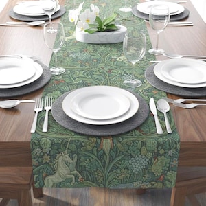 May include: A dining table set for a meal, featuring white plates, silverware, and wine glasses. A green patterned table runner with a unicorn design runs down the center. A small marble planter with white orchids sits in the center.