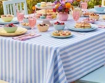 May include: A beautifully set outdoor table with a light blue and white striped tablecloth. The table is laden with colorful food, drinks, and flowers. There are bowls of berries, pastries, and a tiered dessert stand with macarons. Pink drinks with straws are in glasses.
