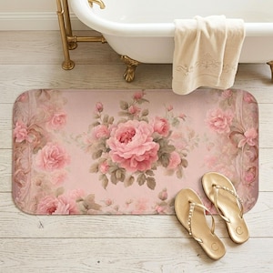 May include: A rectangular bath mat featuring a soft pink background with a central cluster of blooming pink roses and green leaves, accented by ornate, faded scrollwork patterns along the edges, paired with a pair of gold metallic flip-flops.