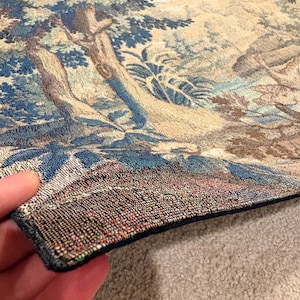May include: A detailed tapestry depicting a landscape scene with trees, foliage, and a river. The color palette includes blues, greens, and beige. The tapestry's edge is visible, showing a dark blue border and intricate stitching.