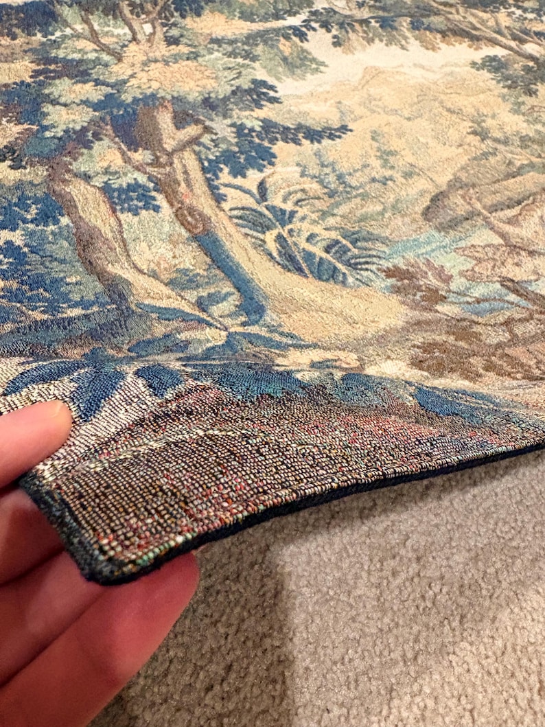 May include: A detailed tapestry depicting a landscape scene with trees, foliage, and a river. The color palette includes blues, greens, and beige. The tapestry's edge is visible, showing a dark blue border and intricate stitching.