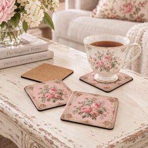 May include: A set of square coasters featuring a pink floral pattern with ornate gold-toned borders and a cork backing. One coaster supports a matching porcelain teacup filled with tea, while two other coasters are displayed on the table surface.